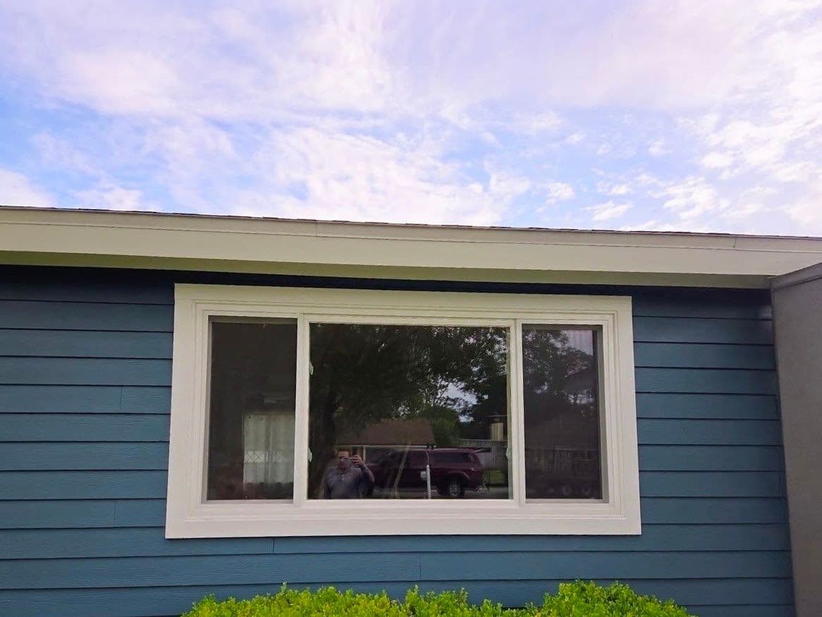Large triple-pane window with white frame installed on blue horizontal siding, reflecting driveway and trees